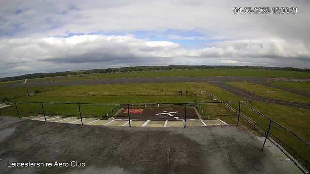 An outdoor view from a webcam at Leicestershire Aero Club shows a cloudy sky with some patches of blue, a grassy field with a fence, a few small paved pathways, and a parking area with white lines marking spaces. In the background, there are open fields and distant hills under a partly cloudy sky. The timestamp in the top right corner indicates the date and time as April 5, 2025, at 3:00 PM.