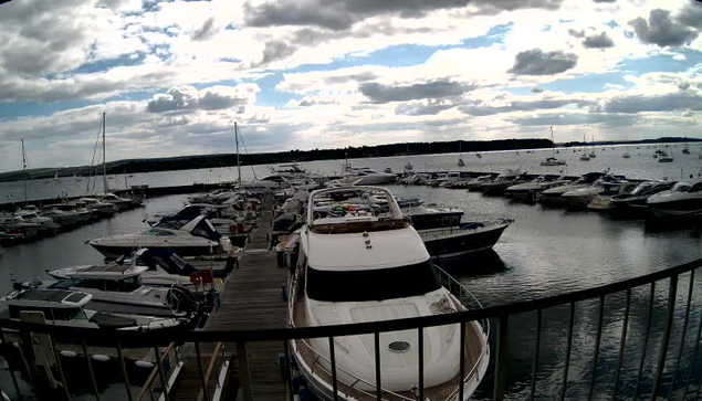 A marina filled with numerous boats and yachts docked along wooden piers, with a cloudy sky overhead and calm water reflecting the vessels.