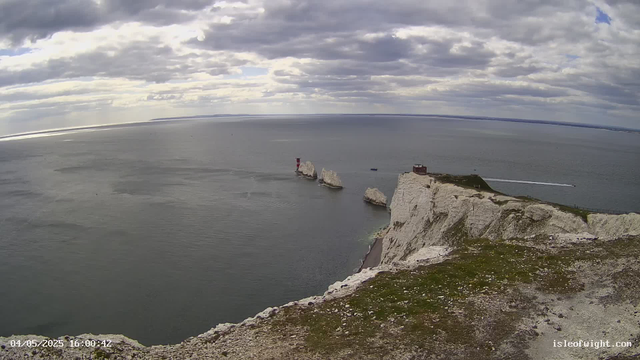 A view from a webcam showing white chalk cliffs along a coastline with the sea extending into the distance. The sky is partly cloudy with patches of sunlight breaking through. A lighthouse stands on a small outcrop among the cliffs, and a few boats are visible near the shoreline. The foreground includes the grassy top of the chalk cliffs with some rocks and dirt.
