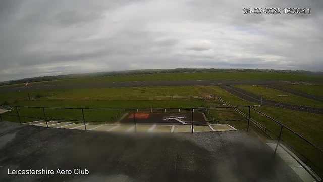 A wide view from a webcam shows an open, grassy airfield with a few small taxiways in the foreground. The sky is overcast with gray clouds, and the landscape stretches out to the horizon with distant trees and fields. There are no aircraft visible on the runway or in the vicinity. The timestamp in the upper right corner indicates the date and time as April 5, 2025, at 16:00:41. The bottom left corner displays the text "Leicestershire Aero Club."