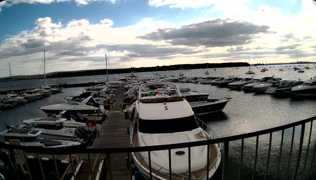 A marina filled with various boats and yachts docked along wooden piers under a partly cloudy sky during sunset. The water is calm, reflecting the boats and the cloudy sky above. A black railing is visible in the foreground.