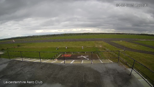 A view from a webcam showing an overcast sky with dark clouds. In the foreground, there is a parking area with several parking spaces and a railing. Beyond the parking lot, a grassy field with a few people standing near a small fenced area and a red platform. The background includes open green fields and a racetrack or paved area extending into the distance. The timestamp in the upper right corner indicates the date and time as April 5, 2025, at 5:00:42 PM. The bottom left corner has the text "Leicestershire Aero Club."