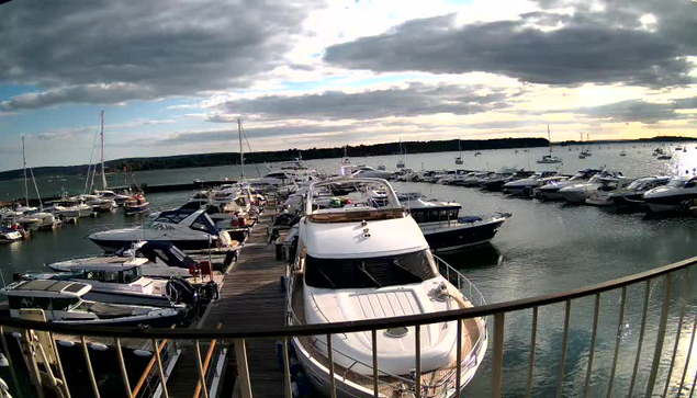 A marina full of boats and yachts docked along wooden piers. The water is calm, and there is a cloudy sky overhead with some sunlight breaking through. The scene is viewed from a balcony or deck with a railing in the foreground.