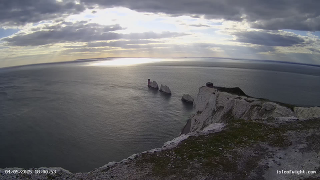 A scenic view of the white chalk cliffs of the Isle of Wight during sunset. The sky is partly cloudy with rays of sunlight breaking through the clouds, reflecting off the calm sea below. The cliffs are rugged and steep, with a small building visible at the top. Three prominent sea stacks rise from the water near the base of the cliffs, with the largest one featuring a lighthouse on top. The date and time stamp at the bottom left indicates "04/05/2025 18:00:53".