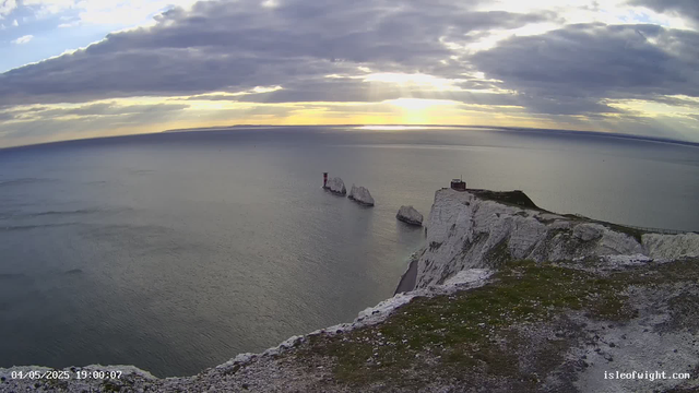 A view of the white chalk cliffs of Dover at sunset, with dramatic cloud cover and rays of sunlight breaking through. The coastline features a small building on top of the cliff and a walking path along the edge. The sea below is calm, and three white chalk stacks are visible in the water near the cliffs. The timestamp shows "04/05/2025 19:00:07" and the website "isleofwight.com" is displayed at the bottom right corner.