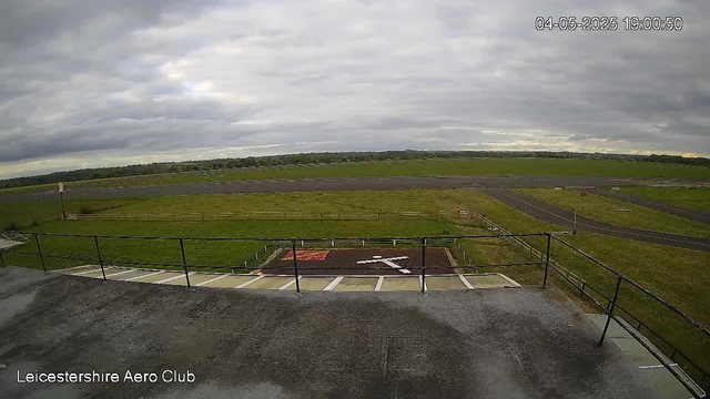 An overcast sky with dark, heavy clouds above a grassy airfield. The view is from a rooftop or high platform, overlooking the airport's runway and taxiways in the distance. There is a parking area with white lines and an arrow on the ground pointing to the right. The scene has a timestamp in the top right corner reading "04-05-2025 19:00:50" and a label in the bottom left corner that says "Leicestershire Aero Club."