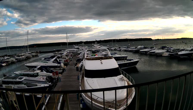 A marina filled with various boats and yachts docked along a wooden pier under a cloudy sky, with some light breaking through in the distance. The foreground shows a black railing, and the water appears calm.
