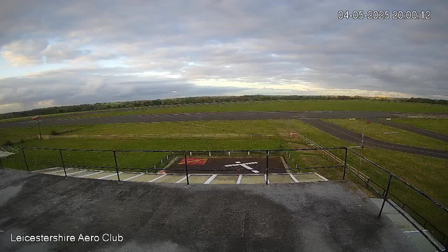 A view from a webcam at Leicesterhire Aero Club showing an open sky with mostly cloudy weather, a runway stretching into the distance, green fields surrounding the airfield, and a helipad painted with a white helicopter symbol in the foreground. The timestamp indicates it was taken on April 5, 2025, at 8:00 PM.