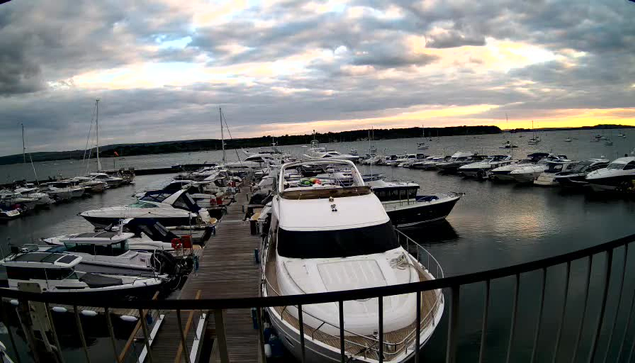 A marina filled with various boats and yachts docked along wooden piers. The sky is cloudy with a hint of sunset near the horizon, casting a soft glow over the water. A black metal railing is visible in the foreground.