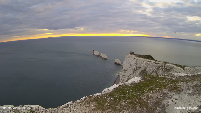 A view of the white chalk cliffs of a coastal landscape at sunset or sunrise, with a lighthouse and small buildings on top of the cliffs. The sea stretches out below, and there are several large rock formations in the water near the base of the cliffs. The sky is partly cloudy with the sun low on the horizon, casting a warm glow.