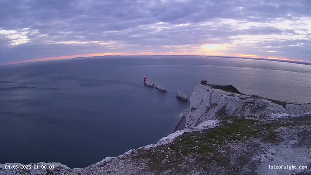 A view of white chalk cliffs by the sea at sunset, with a lighthouse illuminated in red on a small rock formation in the water. The sky is partly cloudy with the sun setting near the horizon, casting a soft glow over the scene.