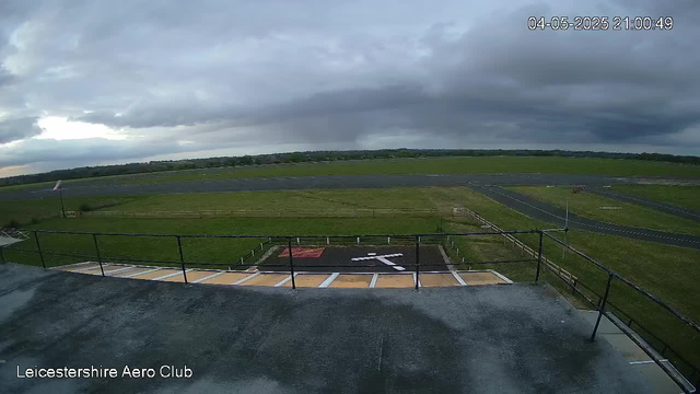 An outdoor view from a webcam shows a cloudy sky over what appears to be an airfield. The foreground includes a concrete area with safety railings and markings painted on the ground. In the background, there are grassy fields and a paved runway stretching into the distance. The timestamp in the top right corner reads "04-05-2025 21:00:49," and the bottom left corner has the text "Leicestershire Aero Club."
