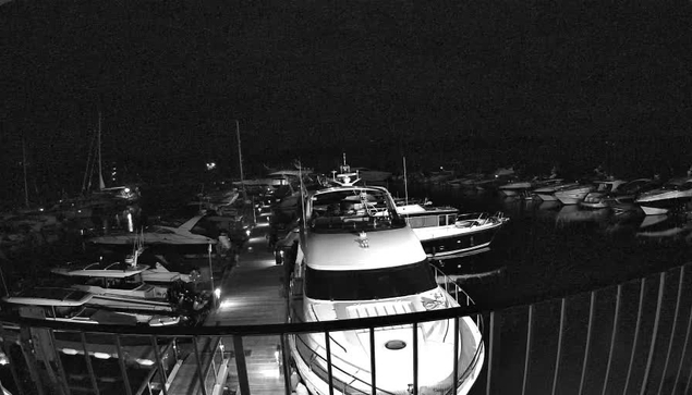 A black-and-white photograph taken from a webcam shows a marina at night. Several boats and yachts are docked closely together along a narrow pier, with more boats visible in the background. The scene is dark, with minimal lighting illuminating the boats and the water. The image is viewable from a high vantage point, possibly from a balcony or a platform, with a curved railing in the foreground.