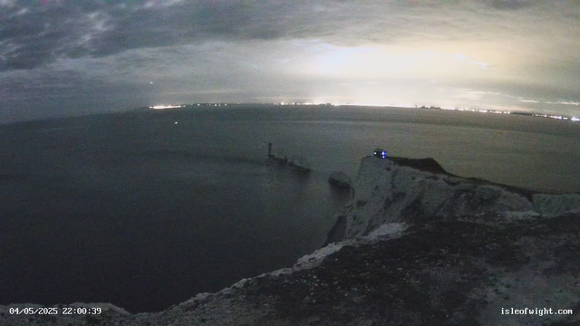 A nighttime scene shows a coastal area with a sea stack or small cliff in the foreground. There is a rocky ledge and some vegetation visible on the right side. In the background, a calm ocean extends toward a horizon lit by distant city lights, with some scattered clouds in the sky. The timestamp indicates it was captured on April 5, 2025, at 22:00.