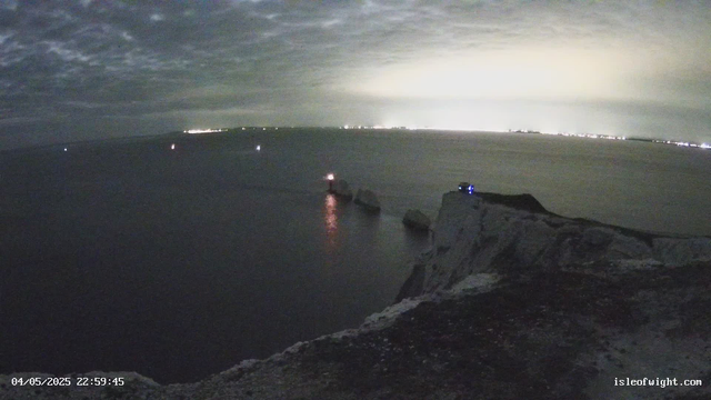 A nighttime view from a webcam overlooking a body of water with a rocky shoreline in the foreground. Several small lights are visible on the water, possibly from boats, and faint city lights are seen on the distant horizon. The sky is cloudy, and there is an ominous glow or lightening in the clouds. The timestamp in the lower left corner reads "04/05/2025 22:59:45," and the website "isleofwight.com" is visible in the bottom right corner.