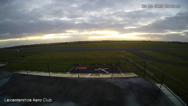A view from a webcam overlooking an open area with a grassy field and a runway. The sky is partly cloudy with the sun either rising or setting near the horizon. There are pathways and a fence around the grassy area, and a small helipad with a large white "H" on it is visible in the foreground. The scene appears calm and quiet, with no people or aircraft present. The timestamp in the top right corner reads "05-05-2025 05:00:58" and the bottom left corner indicates it is from the "Leicestershire Aero Club."