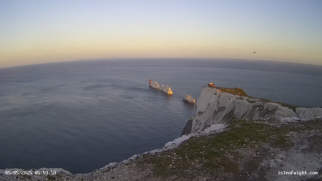 A view from a webcam showing white chalk cliffs with a small building on top, overlooking the sea at sunrise or sunset. Four large rock formations are visible in the water nearby, and a small boat can be seen in the distance. The sky is clear with a faint gradient of color near the horizon. The date and time stamp in the bottom left corner reads "05/05/2025 05:59:58."