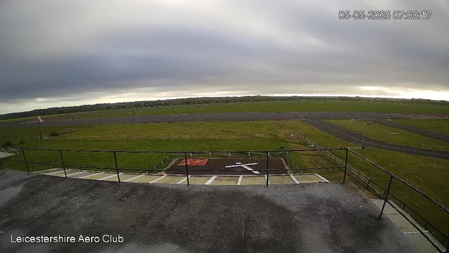 A wide view of an empty airfield on a cloudy day, with grassy runways and taxiways stretching into the distance. The photo is taken from a balcony or platform with a metal railing in the foreground. There's a small area with a red mat or sign near the bottom center. Overcast skies dominate the scene, and the time and date are displayed in the top right corner as "05-05-2025 07:00:17". The bottom left corner contains the text "Leicestershire Aero Club".
