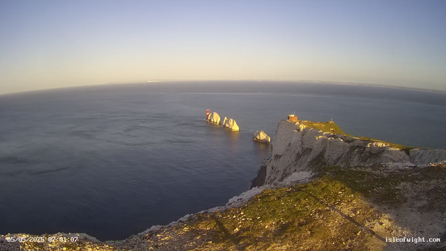 Aerial view of white chalk cliffs overlooking the ocean at sunrise or sunset. Prominent rock formations are visible in the water near the cliffs, with a small building or lighthouse near the edge of the cliff. The sky is clear with a subtle gradient from light to darker blue, and the sea appears calm. The timestamp on the image indicates the date and time as May 5, 2025, at 07:01:07.