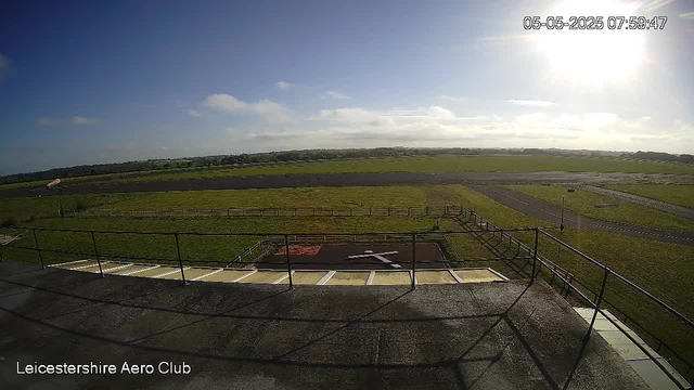 An outdoor view of an airfield taken from a webcam. The image shows a clear sky with a few scattered clouds and the sun shining brightly from the upper right corner. In the foreground, there is a fenced platform or balcony area, and beyond that, open grassy fields stretch out towards the horizon. A small paved runway or taxiway is visible in the middle distance, with a winding road or pathway to the right. The timestamp in the top right corner indicates the date and time as May 5, 2025, at 07:59:47. The bottom left corner has the text "Leicestershire Aero Club."