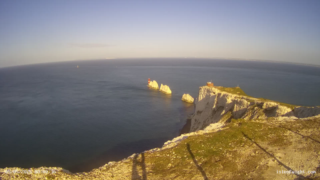 Aerial view of white chalk cliffs with green grass on top overlooking the ocean. Several large white rock formations are situated in the water near the base of the cliffs, with a red and white lighthouse standing on one of the smaller rocks. The sky is clear with the sun casting a warm glow on the cliffs and water.