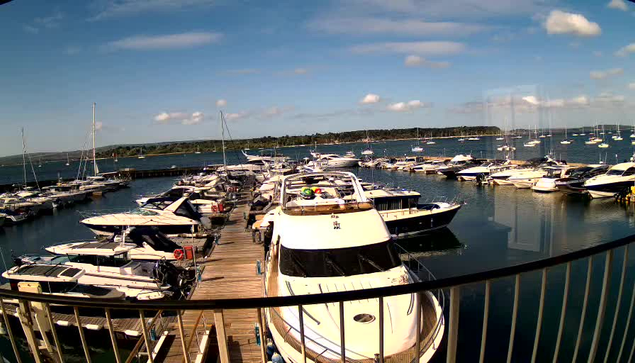 A marina filled with numerous docked boats and yachts, with a wooden pier extending into calm water. The sky is partly cloudy with a few haze clouds, and the horizon shows a tree-lined shoreline in the distance. A curved railing is in the foreground.