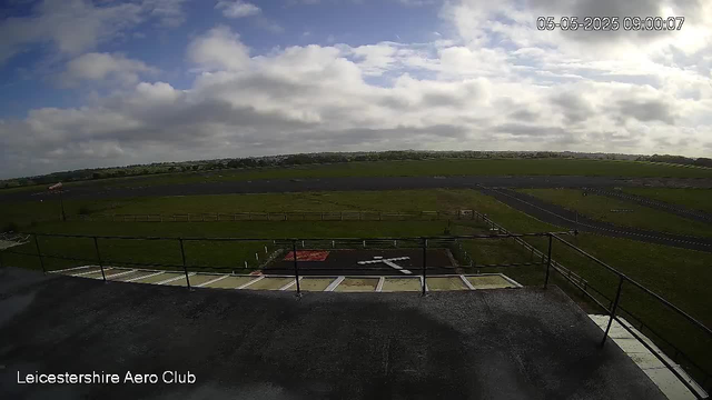 A wide view of an open grassy field with a racetrack running through it under a partly cloudy sky. In the foreground, there is a viewing platform with a black railing and marked parking spots below. The landscape stretches to the horizon with a few distant trees and buildings. The date and time stamp in the upper right corner indicates "05-05-2025 09:00:07."