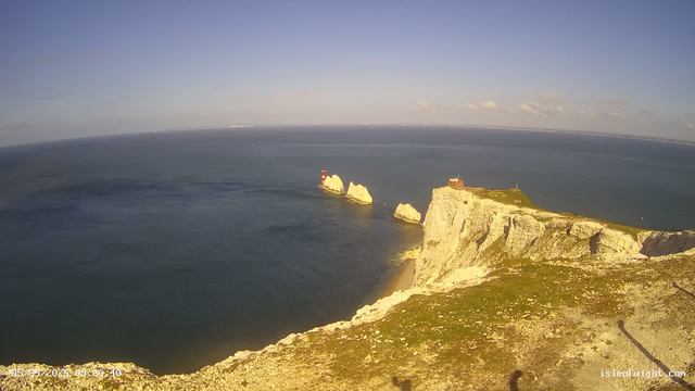 A view from a webcam showing white chalk cliffs along the coastline, with a lighthouse on a small island in the water and another building on top of the cliffs. The ocean is calm and stretches to the horizon under a partly cloudy sky.