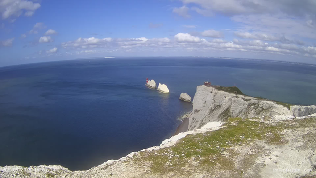 Aerial view of white chalk cliffs along a coastline with a lighthouse on one of the rocks and a building on top of the cliffs. The sea is calm and blue, extending to the horizon under a partly cloudy sky.