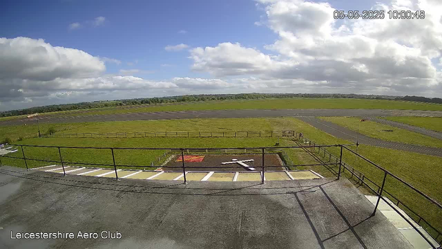 An outdoor view from a webcam at Leicestershire Aero Club showing a clear, partly cloudy sky with a mix of white and gray clouds and patches of blue sky. The scene includes a grassy airfield with a tarmac runway stretching into the distance, surrounded by well-maintained green fields and a few small structures. In the foreground, there is a parking area with white and green markings, as well as a safety railing. The timestamp at the top right indicates the date and time as May 5, 2025, at 10:00:48.