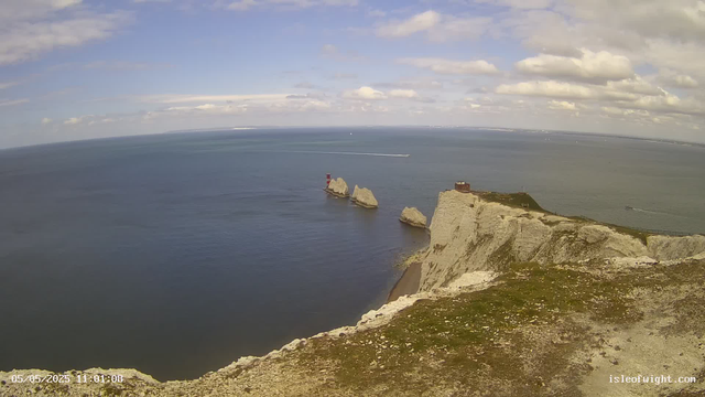 A panoramic view of white chalk cliffs at the edge of the ocean, with a lighthouse on one of the cliffs and several large rock formations in the water nearby. The sky is partly cloudy with patches of blue, and the sea appears calm. In the background, a boat is visible moving across the water. The photo seems to be taken from a high vantage point, possibly from the top of the cliffs. The timestamp on the image indicates it was captured on May 5, 2025, at 11:01 AM.