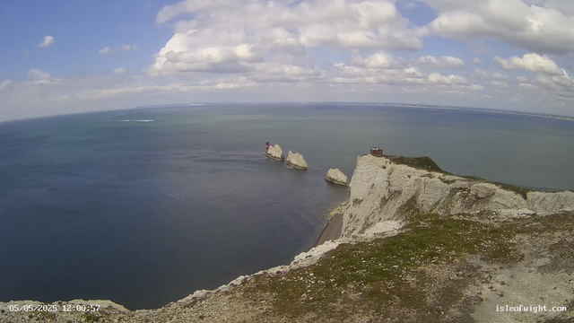 A view from a high vantage point showing white chalk cliffs extending into the sea, with three large rock formations near the shoreline. On top of the cliffs, there is a small building and a path or fence running along the edge. The ocean below is calm, and the sky above is partly cloudy with scattered white clouds. The image has a timestamp in the bottom left corner: "05/05/2025 12:00:57" and a website URL in the bottom right corner: "isleofwight.com".
