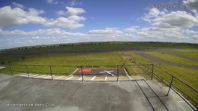 An outdoor view from a webcam showing a bright, partly cloudy sky over a grassy field. In the foreground, there is a small, flat concrete area with parking spaces and a safety railing. Beyond the railing, there are open grassy areas, a tarmac runways or taxiways, and some fencing. A red and white windsock is visible on the left side, indicating wind direction. The image has a timestamp in the upper right corner showing the date and time as 05-05-2025 at 12:00:52, and the bottom left corner displays the text "Leicestershire Aero Club."