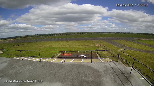 A view from a webcam showing an open outdoor area at Leicestershire Aero Club. The foreground features a concrete surface with a railing, and a designated helicopter landing pad marked with a white H and an arrow. Beyond the landing pad, there is a grassy field with a few small structures and pathways. The background includes a vast green landscape extending to the horizon, under a sky filled with large, fluffy white clouds and patches of blue. The timestamp in the top right corner indicates the date and time as 05-05-2025 at 12:59:53.