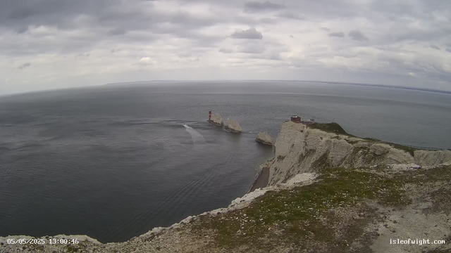 A view from a high vantage point overlooking white chalk cliffs bordering the sea. In the distance, there are rock formations in the water, with one featuring a small red and white lighthouse on top. The sea is calm with gentle ripples, and the sky is overcast with thick gray clouds. The timestamp in the bottom left corner credits the date as May 5, 2025, and the time as 13:00:46. The bottom right corner displays the website isleofwight.com.