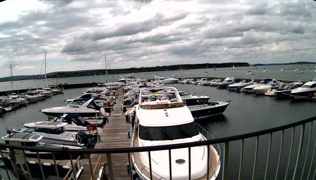 A marina filled with numerous boats and yachts docked along wooden piers. The water is calm, and the sky is overcast with dark clouds. There is a black metal railing in the foreground, curving along the edge of the dock. In the background, a distant shoreline with trees can be seen.