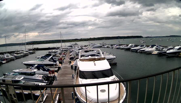 A marina filled with numerous boats and yachts docked along a wooden pier, with water on both sides. The sky is cloudy with dark, overcast clouds, and a distant shoreline is visible on the horizon.