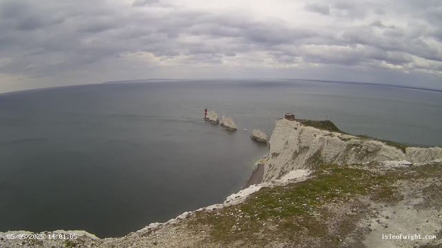A view of white chalk cliffs along the coastline with a small building on top of the cliffs. In the water below, there are three large white chalk sea stacks, with one having a red navigational marker on top. The sky is cloudy with a mix of dark and light gray clouds. The water appears calm, and the horizon extends into the distance. The scene captures a dramatic coastal landscape under overcast skies.