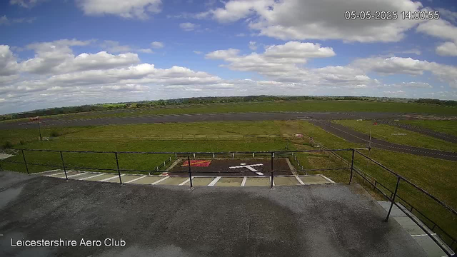 A view from a webcam at Leicesterhire Aero Club shows a partly cloudy sky with scattered white clouds over green fields and a distant horizon. In the foreground, there is a platform with black safety railings, and below it, several bicycle parking spaces and a small red and white sign. The timestamp on the image reads 05-05-2025 14:00:55.