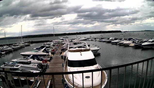 A marina with numerous boats docked along wooden piers extends into a body of water. The boats are mostly white and vary in size, with some larger yachts in the foreground. The sky above is filled with dark, heavy clouds, suggesting overcast weather or an approaching storm. A black metal railing is visible in the foreground, indicating the photo was taken from a platform or walkway overlooking the marina.