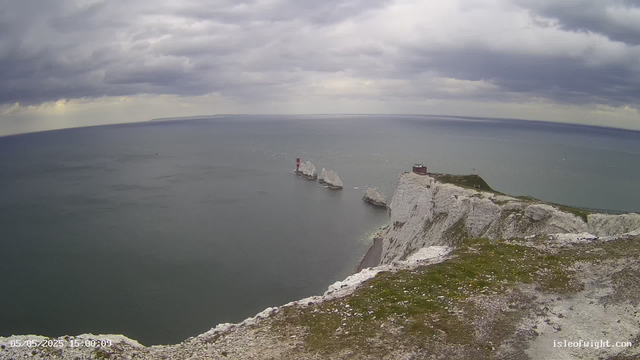 A scenic view of white chalk cliffs overlooking the ocean on a cloudy day. There are three large rock formations emerging from the water near the cliffs, and a small building or lighthouse is situated on top of the highest point of the cliffs. The sky is filled with dark, heavy clouds, suggesting an overcast weather. The photo is dated May 5, 2025.