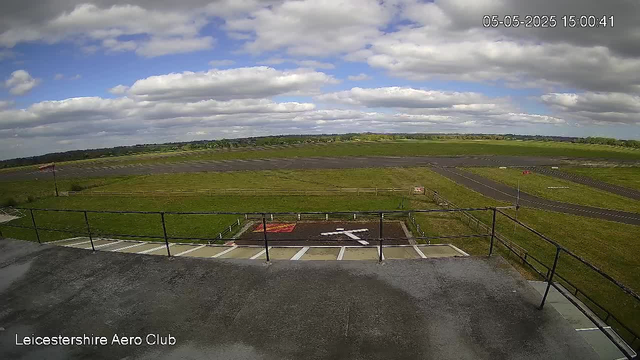 A wide view of an airfield shows a grassy landscape with a runway in the distance, under a partly cloudy sky with patches of blue. In the foreground, there is a concrete parking area with reserved parking spaces and a black railing, and an area marked with a large white arrow pointing left. The image includes a timestamp in the top right corner reading "05-05-2025 15:00:41" and a label in the bottom left indicating "Leicestershire Aero Club."