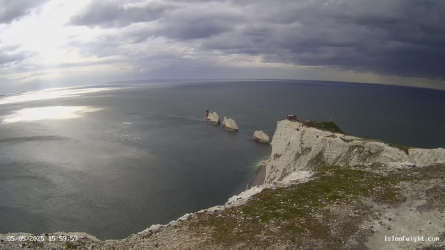 A view of white chalk cliffs and the ocean beneath a cloudy sky. The cliffs are rugged and steep, with some patches of grass on top. In the distance, there are large rock formations in the water, one of which has a small lighthouse on it. The scene is captured during daytime with sunlight reflecting off the water.