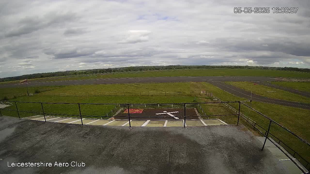 An outdoor view from a webcam at Leicesterhire Aero Club showing a cloudy sky with patches of light breaking through. In the foreground, there is a small parking area with white lines marking spaces, bordered by a black metal railing. Beyond the parking area, there is a grassy open space with a few small fences. A runway extends into the distance, and a small flag is visible on a pole on the left side of the image. The timestamp on the top right corner reads "05-05-2025 16:00:07."