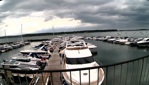 A marina filled with numerous docked boats and yachts under a cloudy sky, with a wooden pier extending into the water and a metal railing in the foreground.