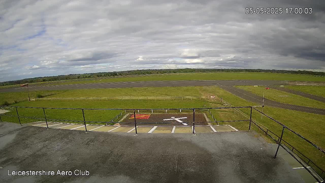 A view from a webcam at LeicesterShire Aero Club showing a large open grassy field with a runway extending across the landscape. The sky is mostly cloudy with patches of sunlight. In the foreground, there is a parking area with marked parking spaces and a white arrow pointing to the right, enclosed by a black metal railing. The timestamp in the top right corner reads May 5, 2025, at 17:00:03.