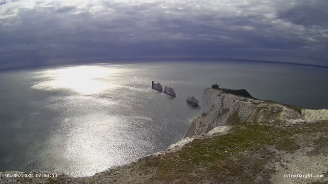 A view from a webcam shows white chalk cliffs overlooking the sea, with several large sea stacks just off the coast. The sky is partly cloudy with the sun reflecting brightly on the water's surface, creating a shimmering effect. The foreground features a grassy, rocky edge of the cliffs. In the distance, the horizon stretches across the ocean. The timestamp on the image reads "05/05/2025 17:00:13" and a footer indicates the source as "isleoftight.com."