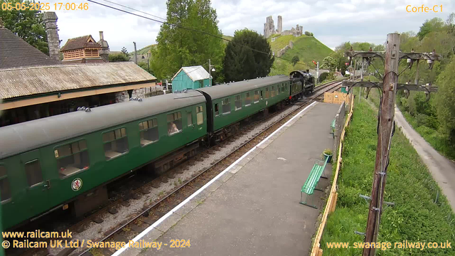 A vintage green train with multiple carriages is stationary on the tracks next to a small station platform. A black steam locomotive is attached at the front. The station has a yellow safety fence, green benches, and some potted plants. Overhead, power lines run alongside the tracks. In the background, there is a hill with the ruins of an old castle and surrounding greenery, including trees and grassy areas. A narrow country road runs parallel to the railway line on the right side of the image. The sky is mostly cloudy.