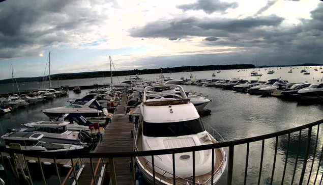 A marina filled with numerous boats and yachts docked along wooden piers. The water is calm, and the sky is partly cloudy with dark clouds overhead, suggesting an approaching storm or late afternoon. The view is from a balcony or platform with a curved railing in the foreground.