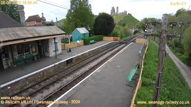 A small railway station platform with two sets of train tracks running alongside it. There are green benches on the platform, a small blue shed, and a yellow fence near the edge. To the right, a wooden utility pole with wires extends over the platform. In the background, there is a green hill with a castle ruin on top and some buildings at the base. Trees with fresh green leaves surround the area, and a narrow dirt path runs parallel to the tracks on the right side. The sky is overcast. The timestamp indicates the date as May 5, 2025, and the time as 18:00:40.
